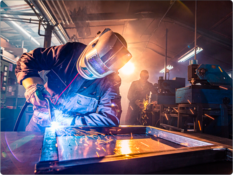 A worker wearing a face shield and gloves bends over his work while welding a steel frame.