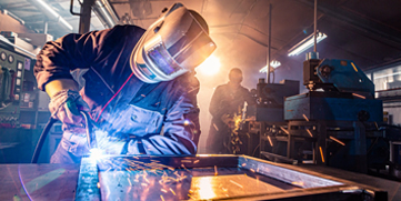 A worker wearing a face shield and gloves bends over his work while welding a steel frame. 